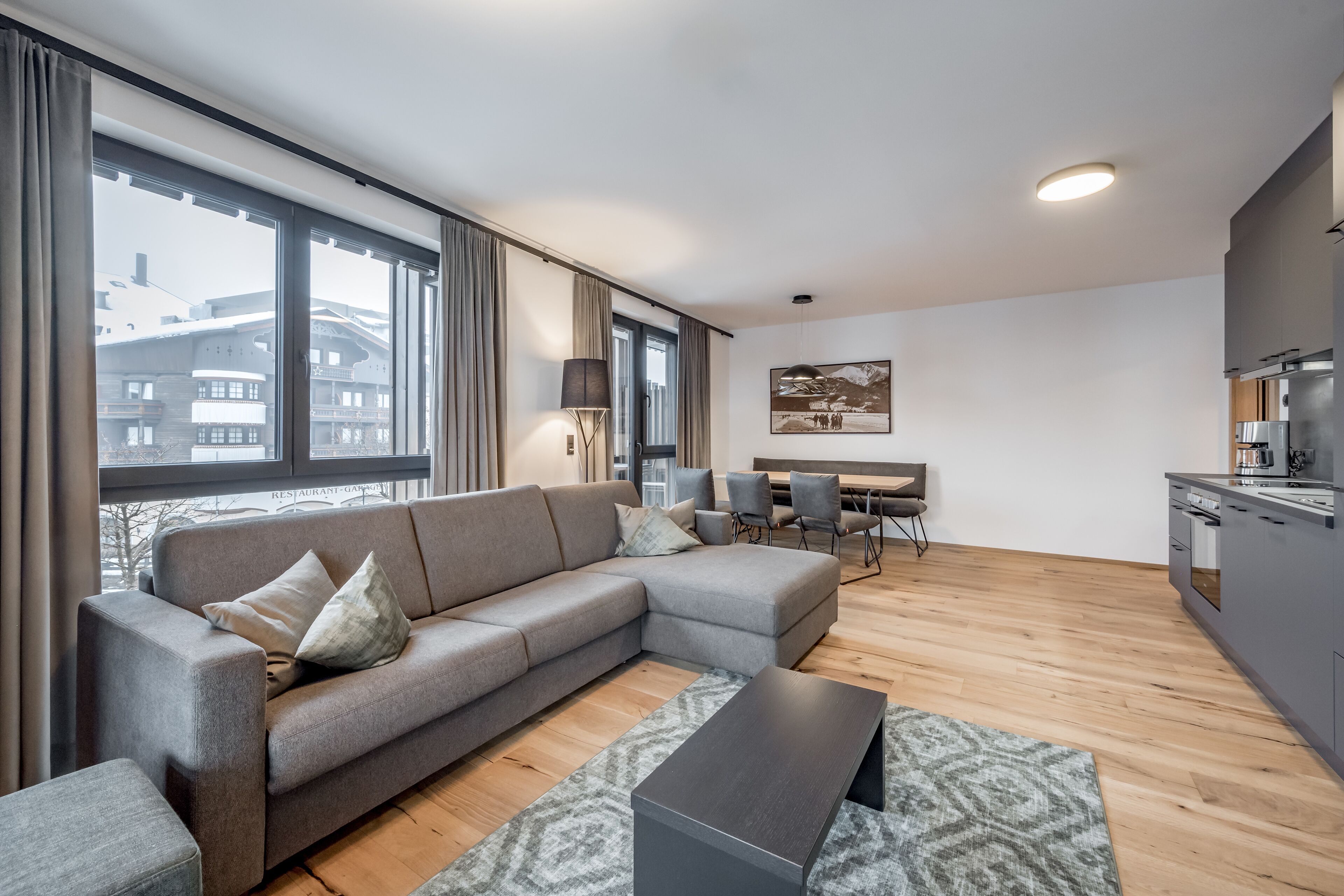 A modern living room with a grey sectional sofa, a small coffee table, and a dining area with chairs and a table. There is a kitchen area with a stove and cabinets. Large windows provide a view of buildings outside.
