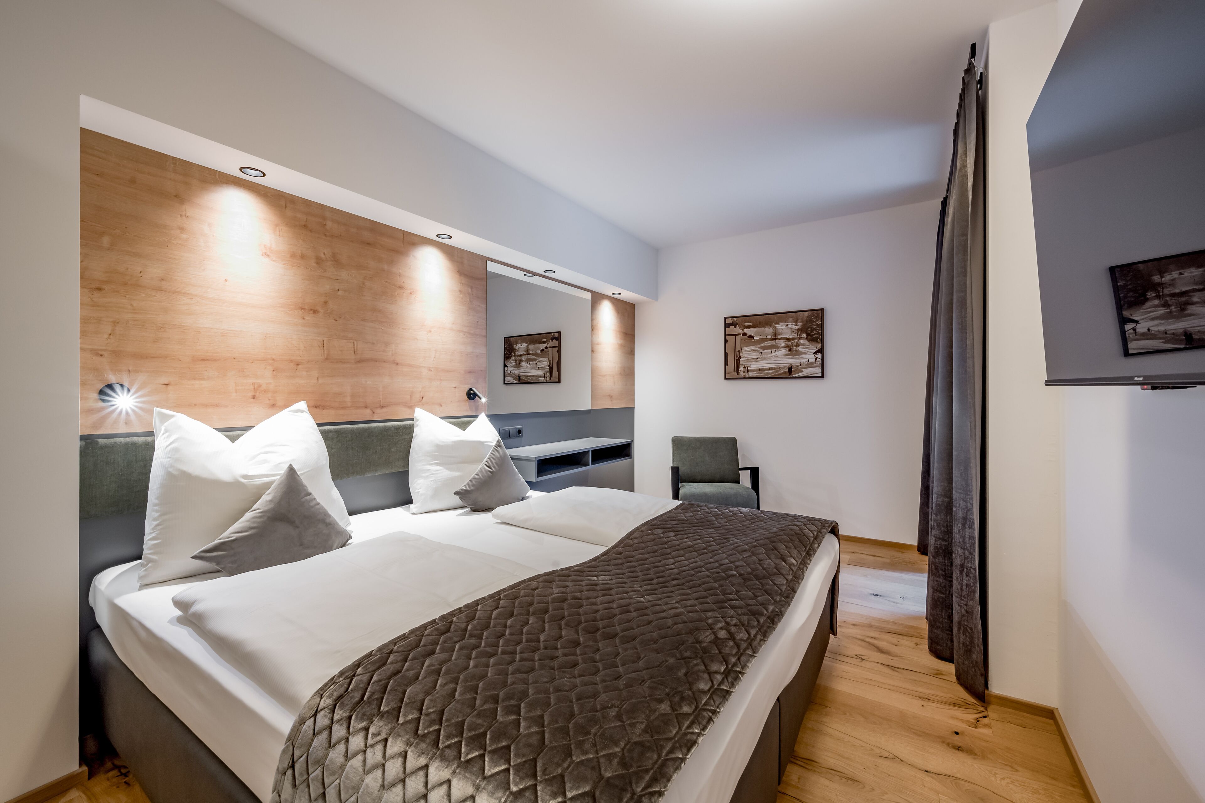 A modern bedroom with a wooden headboard, a bed with white linens and a textured blanket, a grey chair, and artwork on the walls.