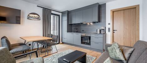 A modern living room with a sofa, coffee table, and TV. A kitchen with grey cabinets and a wooden door is visible.