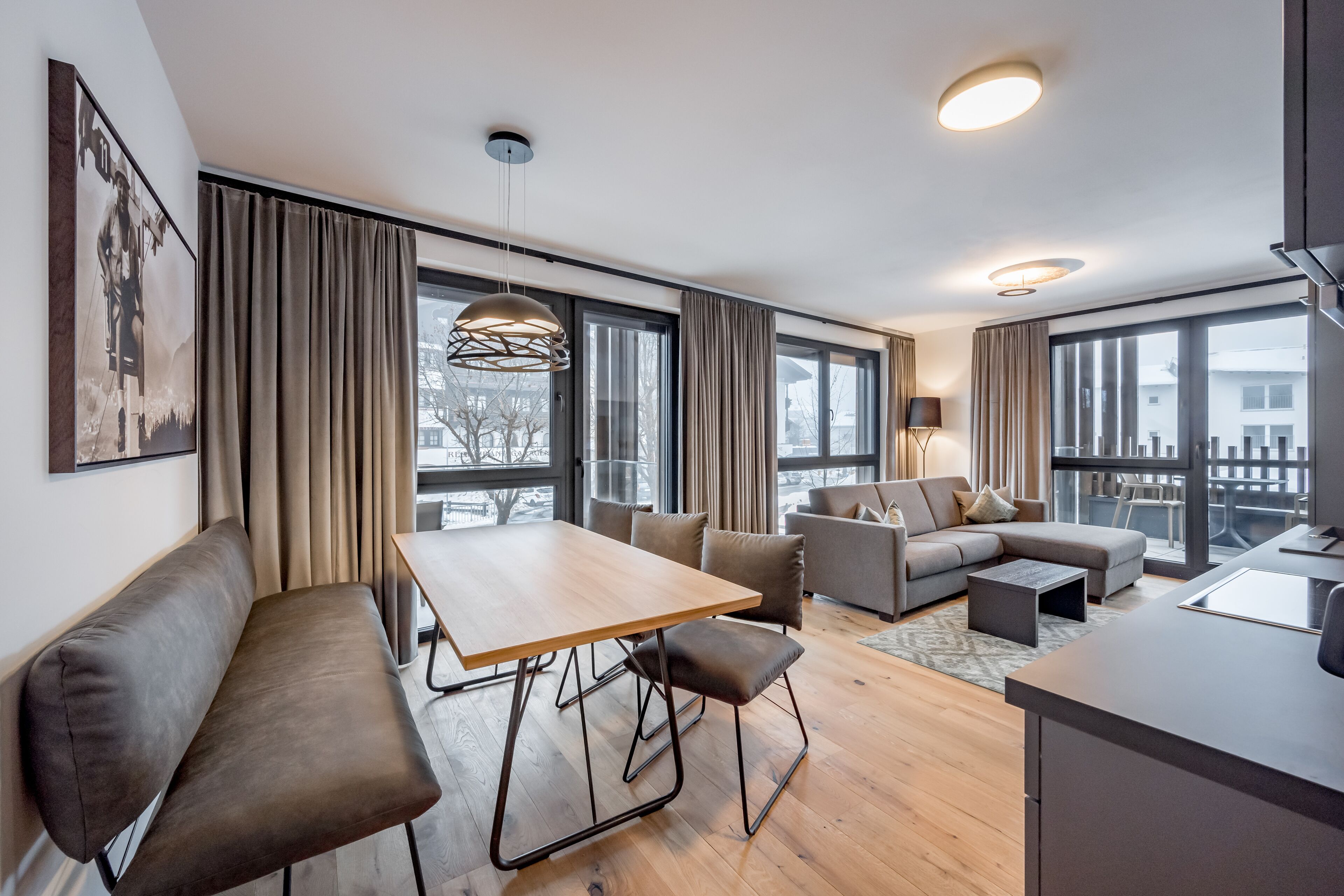 Ett modernt vardagsrum med ett matbord i trä, en soffa och en bänk. Det finns stora fönster med gardiner, en hängande lampa och utsikt över omgivningen.