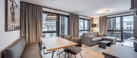 Ett modernt vardagsrum med ett matbord i trä, en soffa och en bänk. Det finns stora fönster med gardiner, en hängande lampa och utsikt över omgivningen.