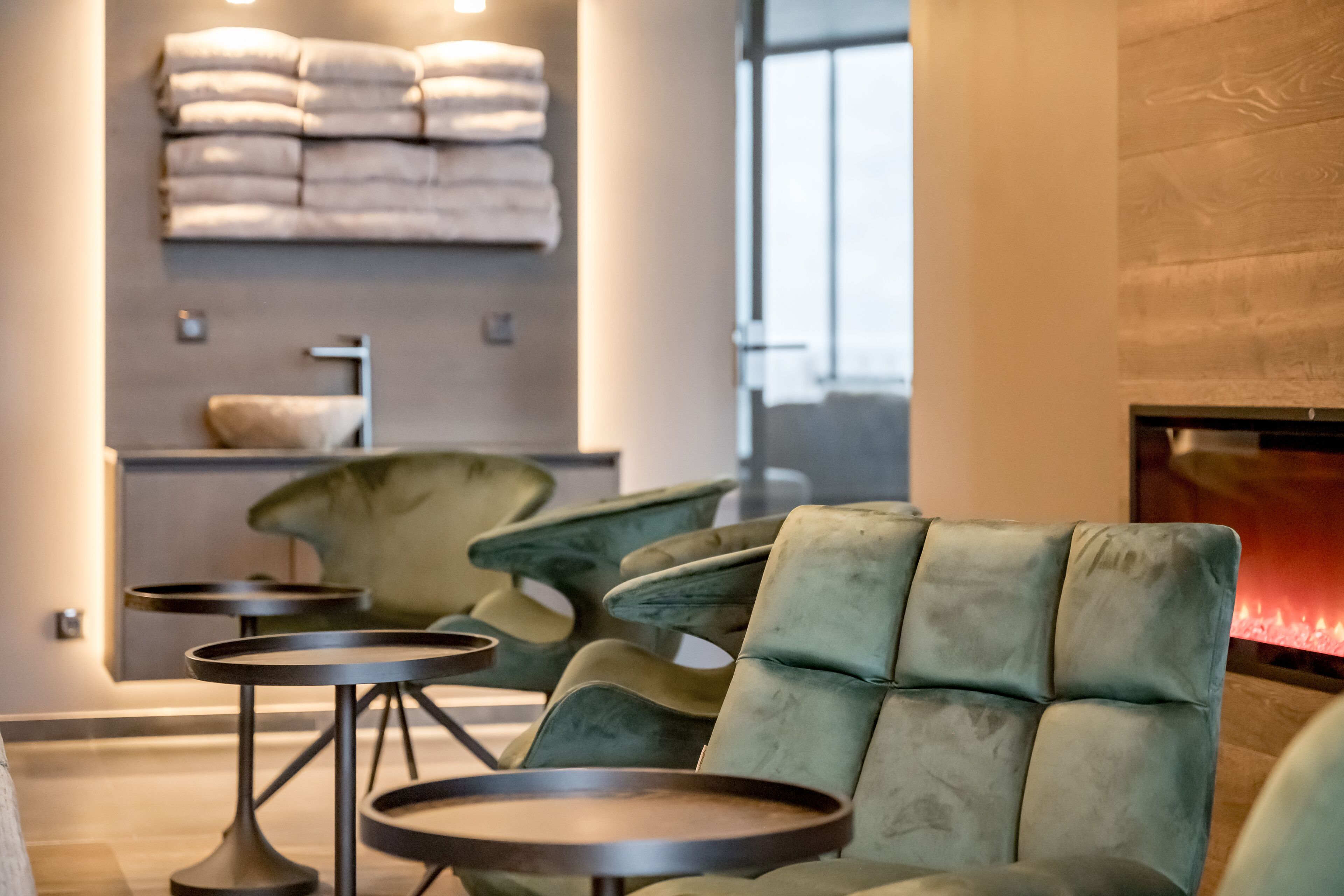 A modern lounge area with green upholstered chairs, small round tables, and a fireplace.