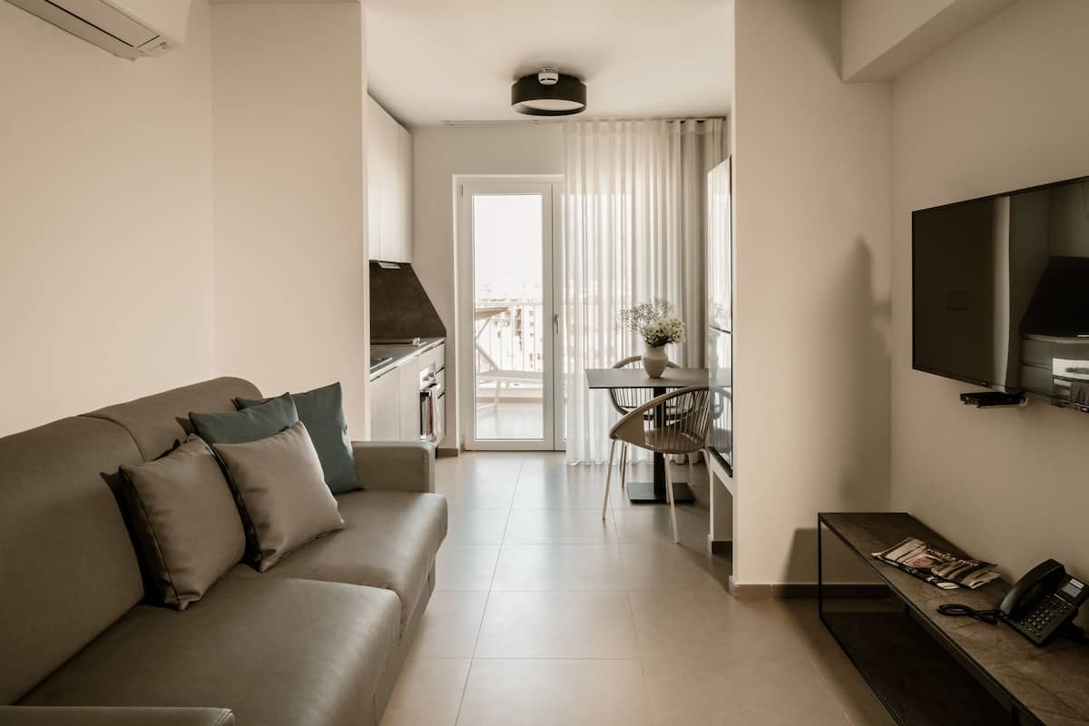 A modern living room with a sofa, a coffee table, and a TV. There is a balcony with a view of buildings.