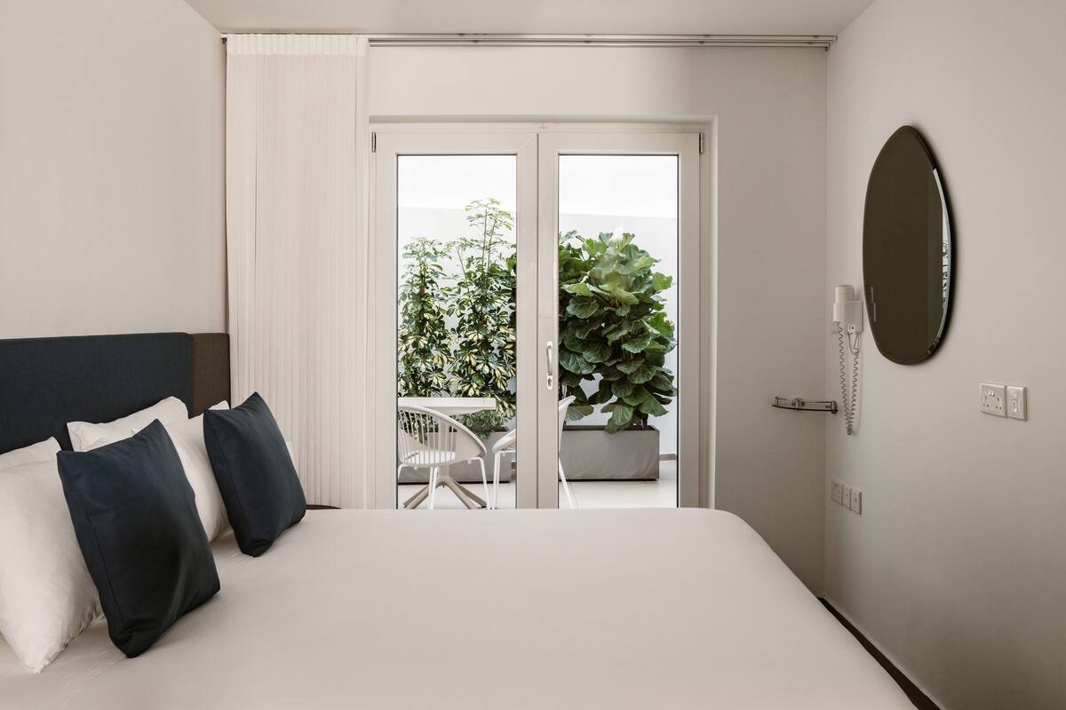 A hotel room with a bed, white curtains, a view of a balcony with plants, and a round mirror on the wall.
