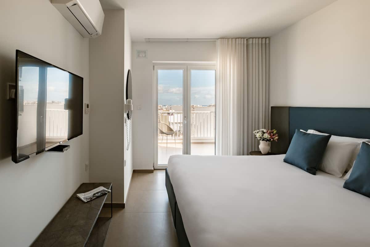 A hotel room with a bed, a TV, a balcony with a view, and a window with curtains.