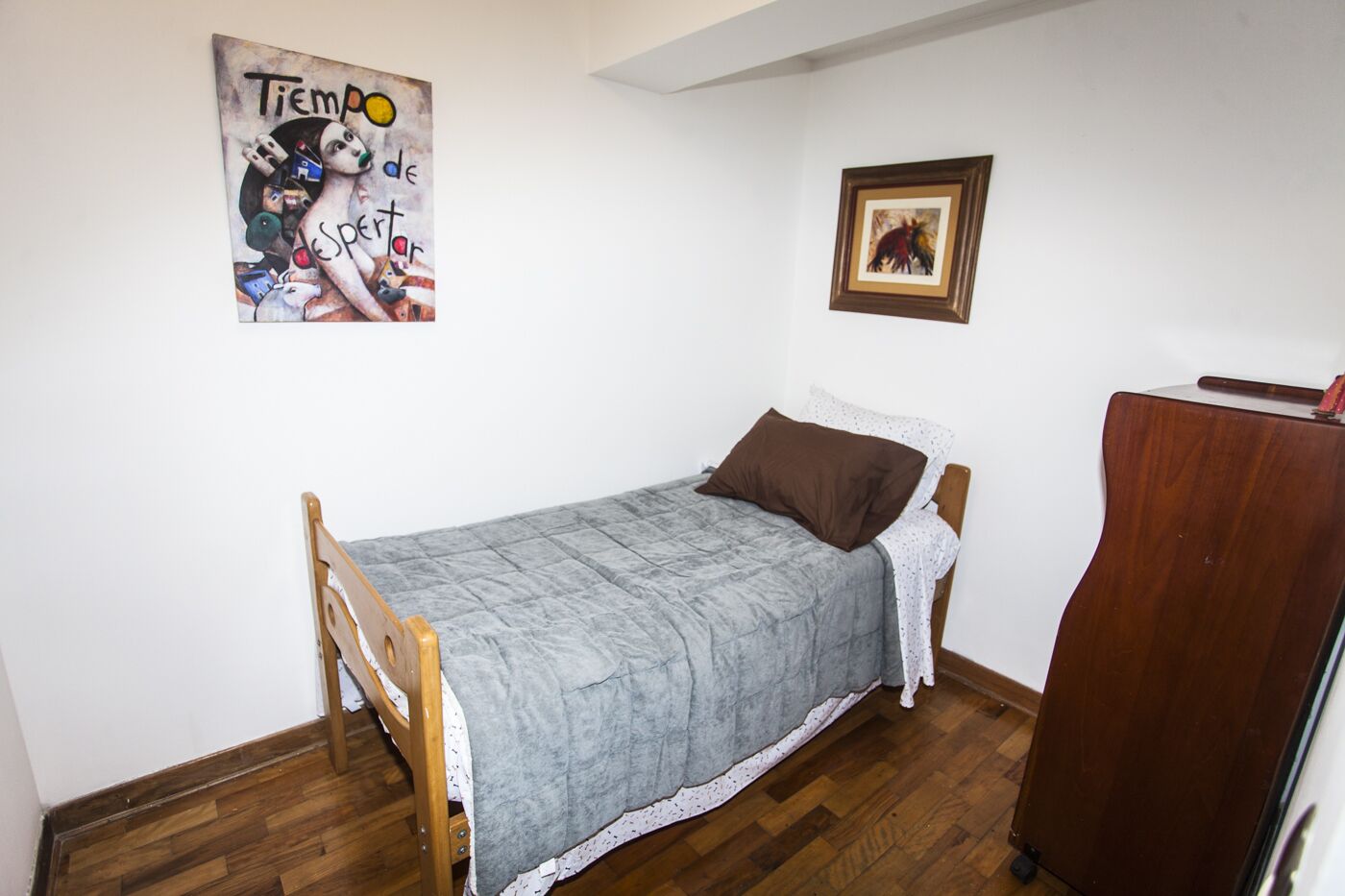 Quarto pequeno com cama de solteiro, estrutura de cama em madeira, colcha cinza e travesseiro marrom. Um guarda-roupa de madeira à direita e um quadro na parede.
