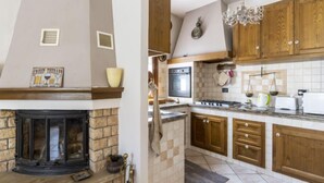 A kitchen with a stone fireplace, wooden cabinets, a stainless steel stove, and a hanging chandelier. - Vacation home Anna by Interhome (Ville San Pietro)