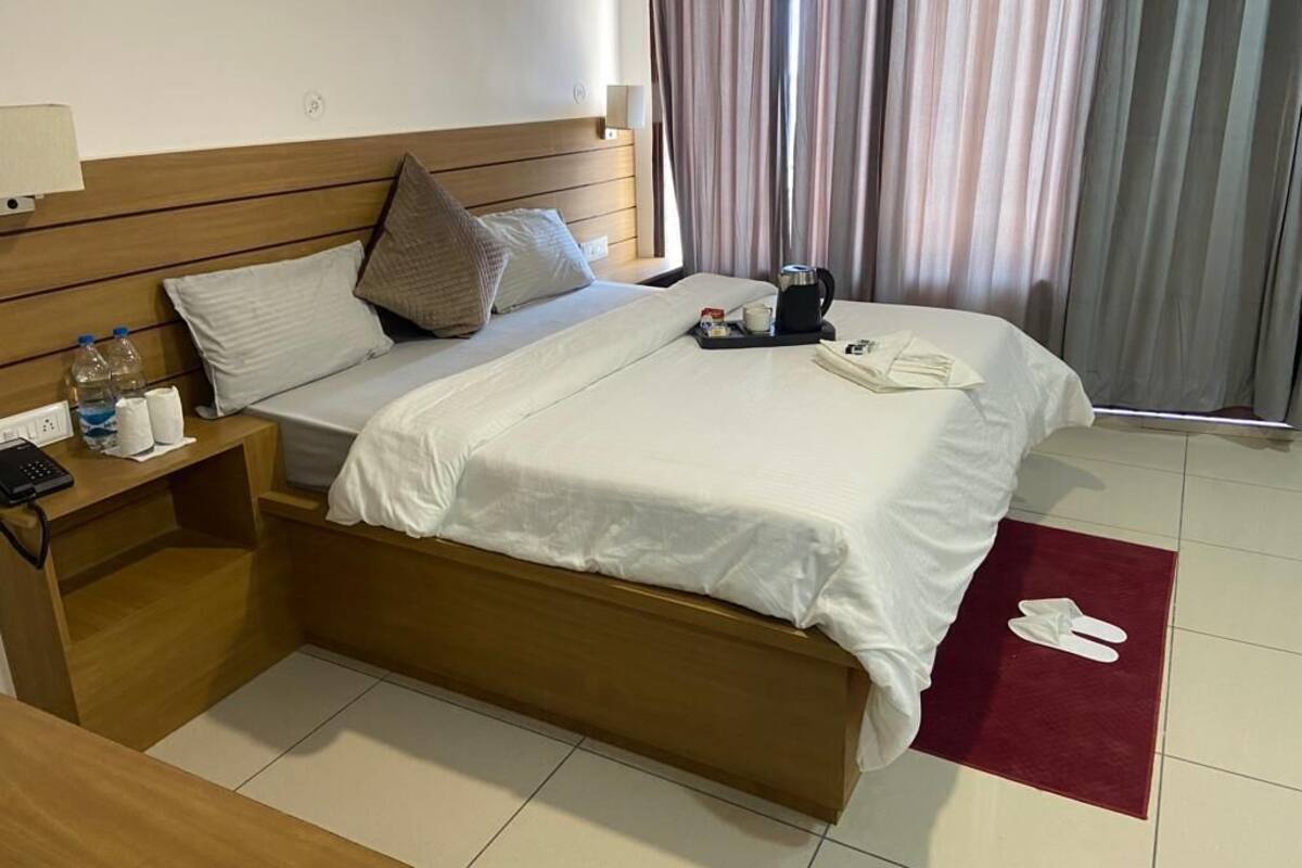 A hotel room with a wooden bed, a nightstand with a lamp, a coffee maker, and a red rug with slippers.