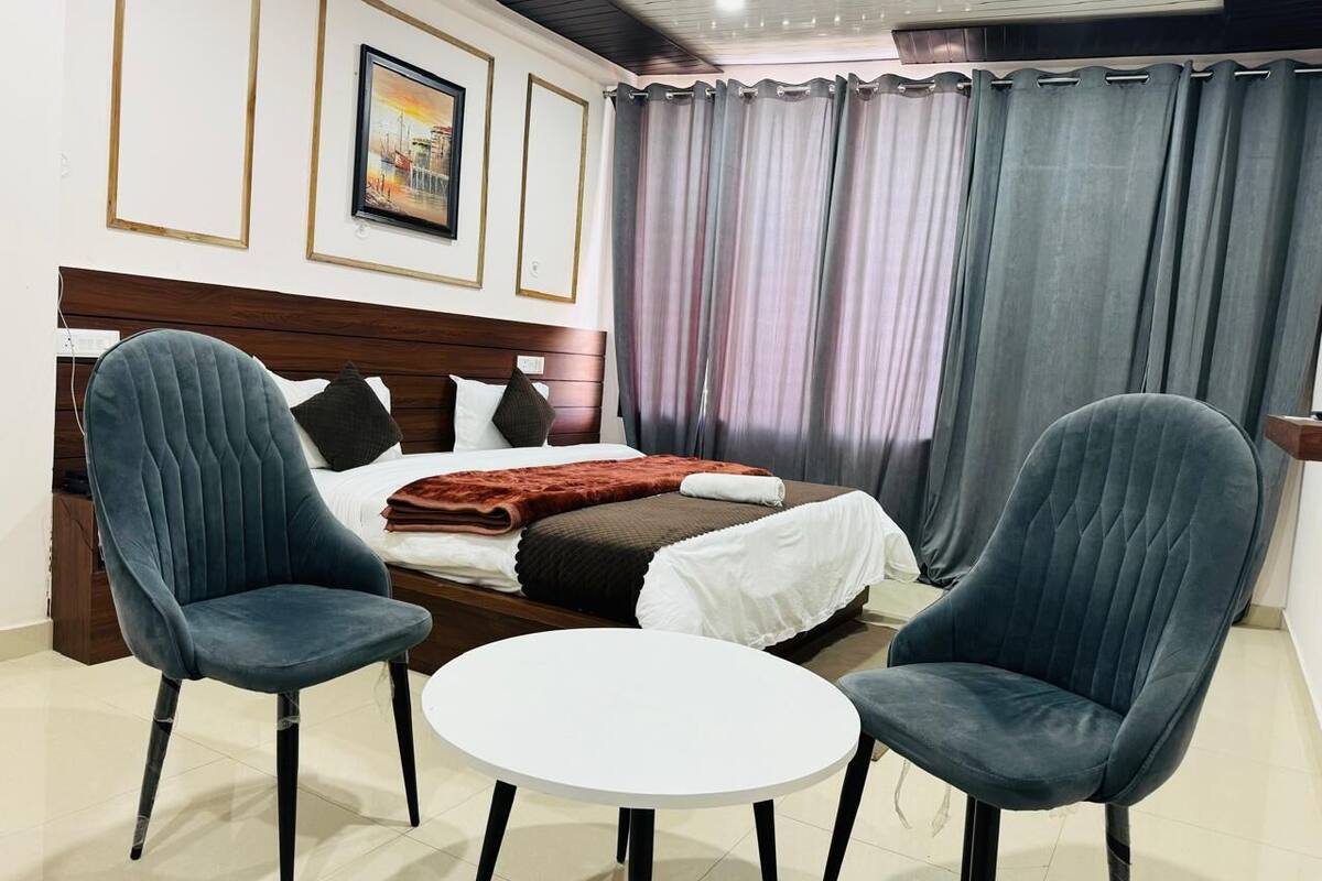 A hotel room with a bed, two armchairs, a small round table, and a view of the city through curtains.
