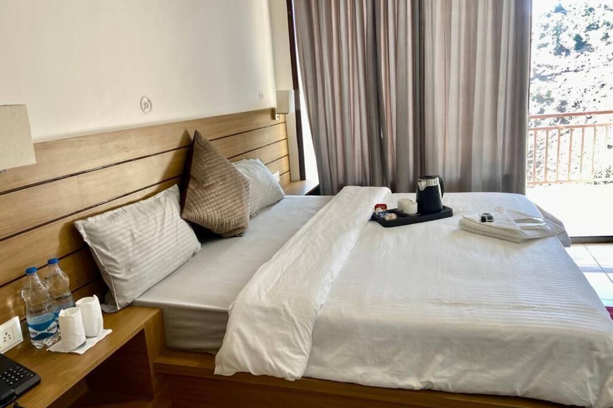 A hotel room with a wooden headboard, a bed with white bedding, a bedside table with a phone and toiletries, and a window with curtains.