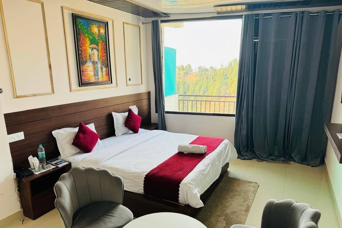 A hotel room with a large bed, two chairs, a small table, a nightstand, a painting on the wall, and a balcony with curtains.