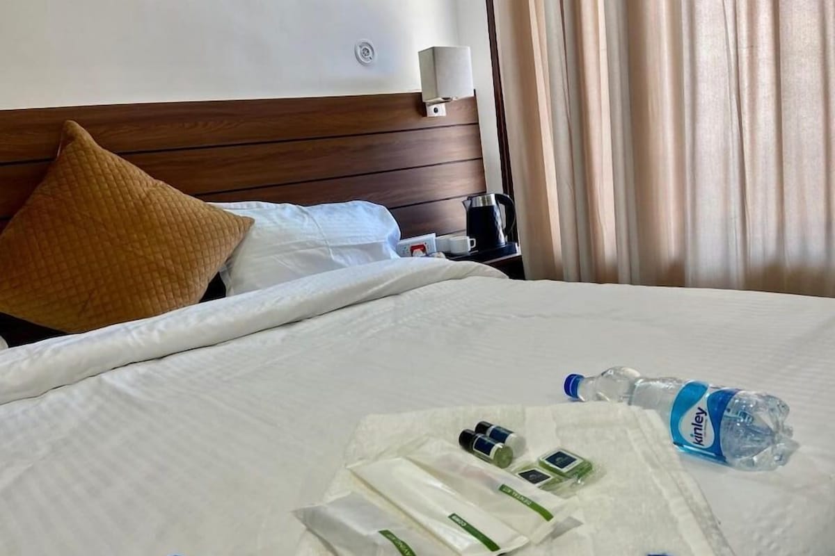 A hotel room with a neatly made bed, a pillow, a bedside table with a lamp, a water bottle, and a tray with toiletries.
