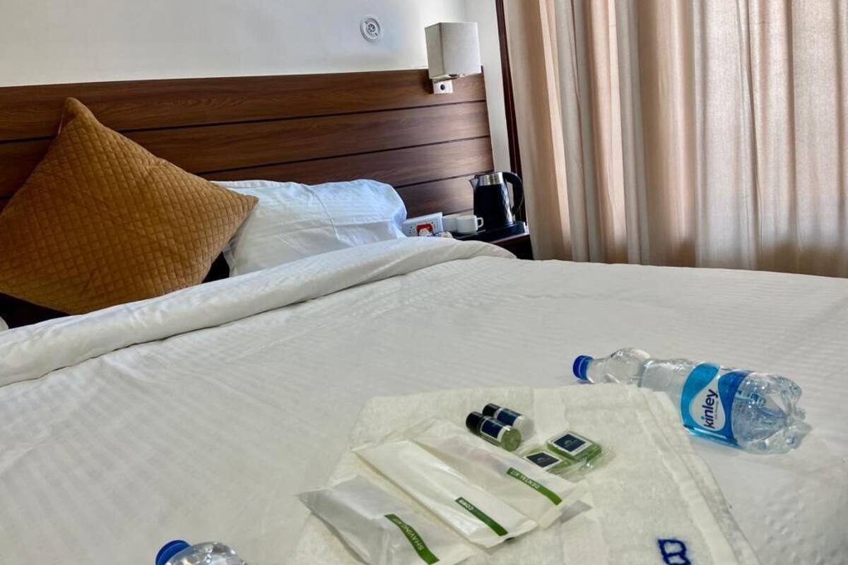 A hotel room with a neatly made bed, a bedside table with a kettle, a bottle of water, and toiletries on the bed.