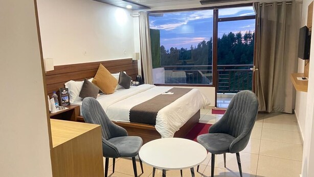Habitación de hotel con cama, dos sillones, una mesita, televisor y balcón con vistas a árboles.