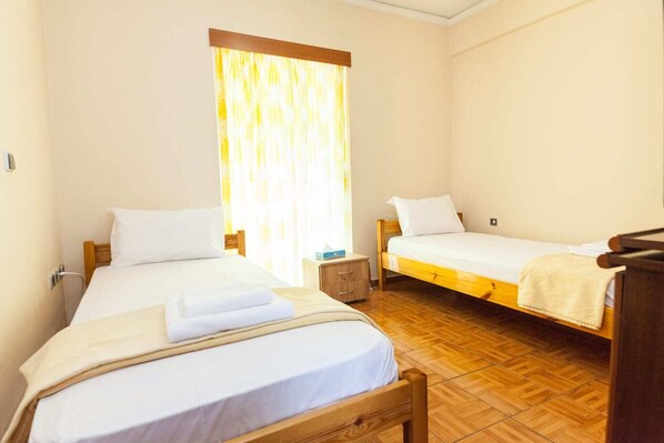 A room with two single beds, a wooden nightstand, a small table, and a window with curtains. - Cute one bedroom apartment in Pireas (Piraeus)