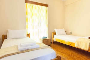 Habitación de hotel con dos camas individuales, cabeceros de madera, mesitas de noche pequeñas y una ventana con cortinas.