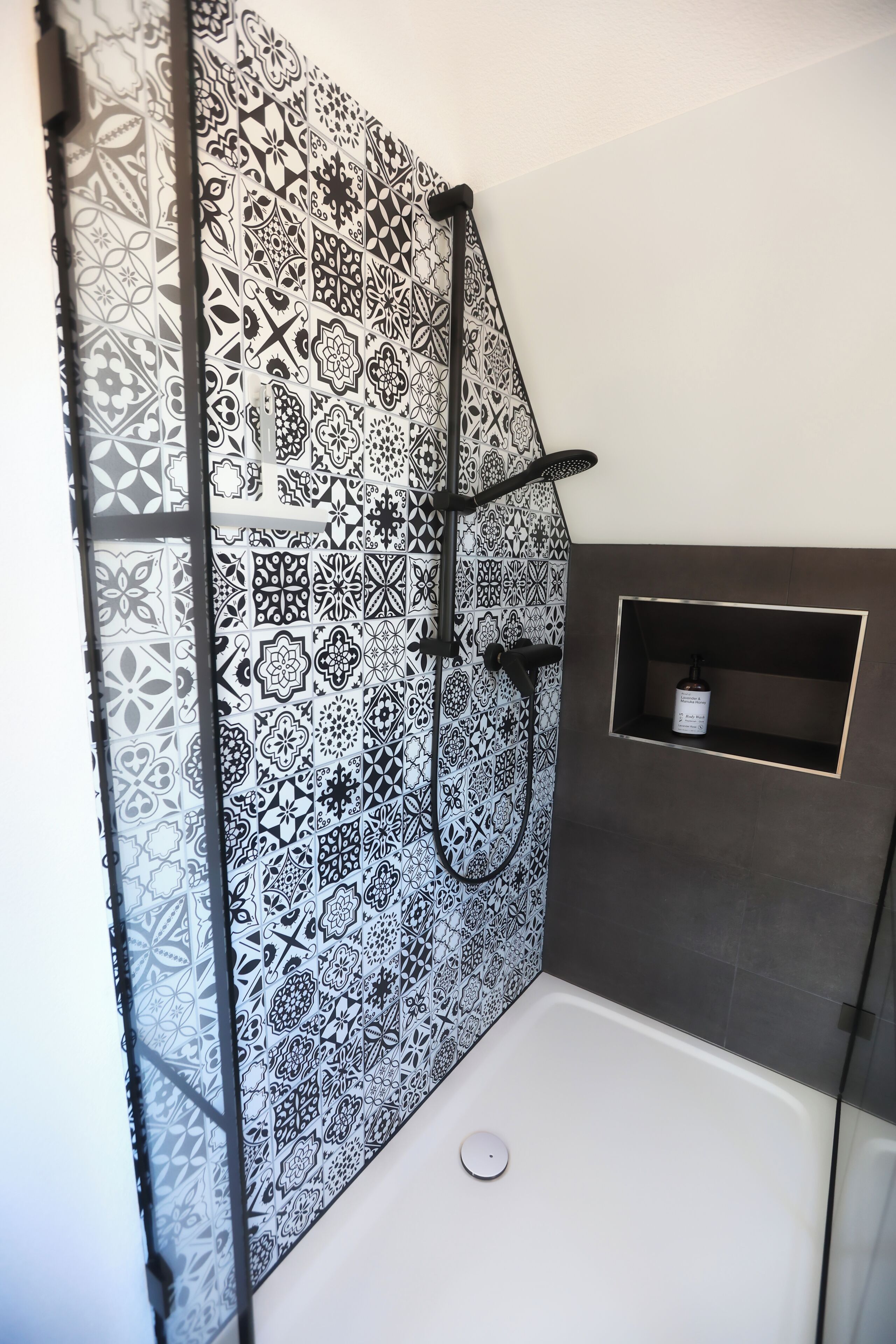A bathroom with a shower area featuring a black and white patterned tile wall, a glass-enclosed shower, and a small shelf with a bottle.
