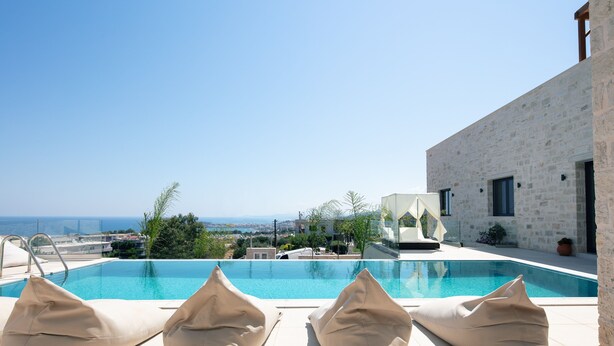 Une piscine à débordement sur le toit, agrémentée de coussins beige, offrant une vue dégagée sur la mer, avec un bâtiment en pierre en arrière-plan.