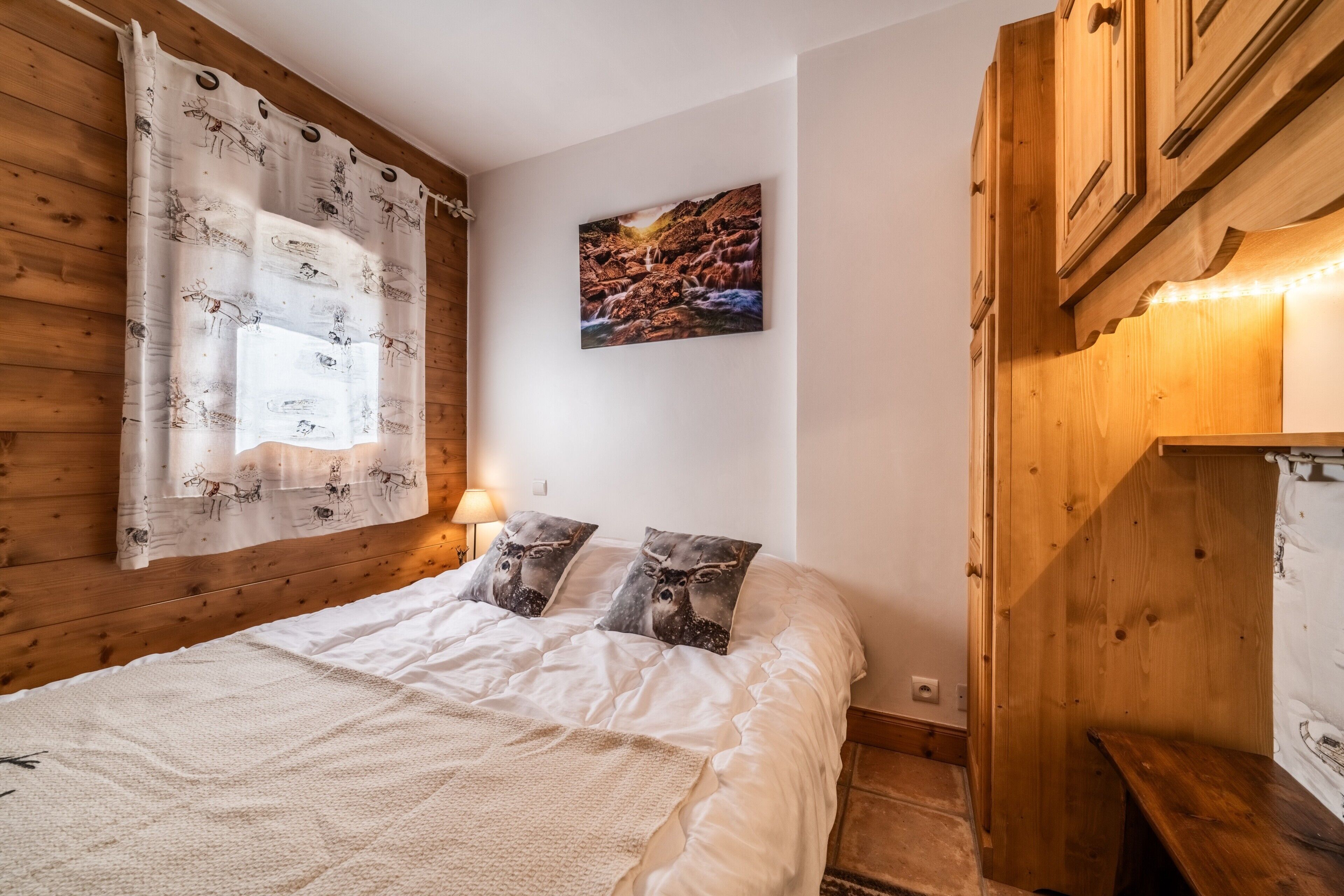 A bedroom with a wooden headboard, a bed with white bedding, two decorative pillows, a wall-mounted painting, and a bedside lamp.