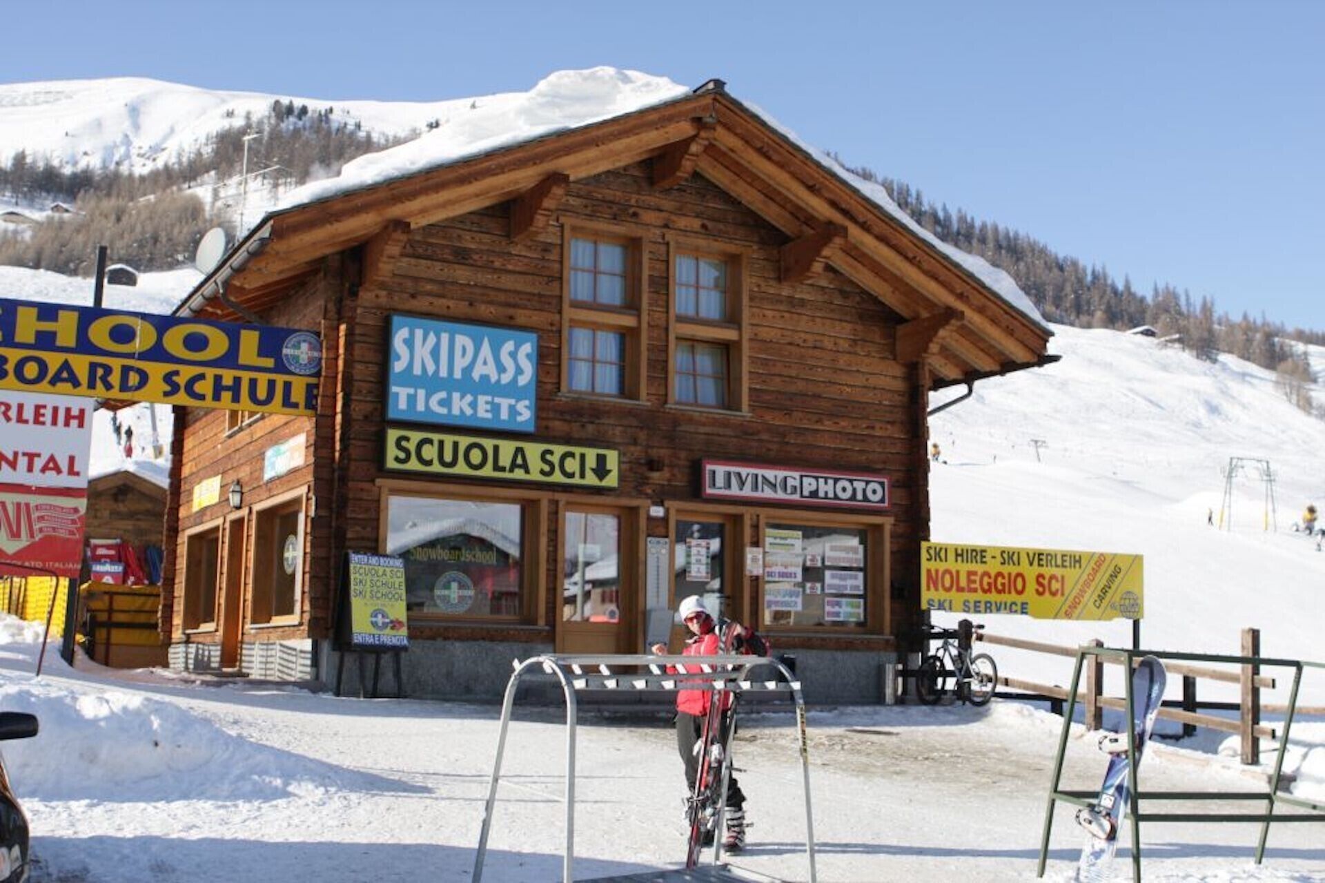 A wooden building with signs for 'Skipass Tickets,' 'Living Photo,' and 'Ski Hire - Ski Verleih.'