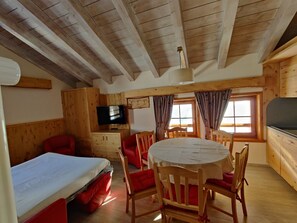 A wooden cabin interior with a bed, a dining table with chairs, a TV, and a window with curtains.