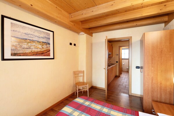 A room with a bed, a chair, a wooden ceiling, a framed painting, and a view into a kitchen area.