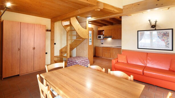 Een houten eettafel met stoelen, een rode bank, een keuken met een magnetron en een trap.