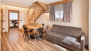 A living room with a wooden staircase, a grey sofa, and a dining table with chairs. - Apartment 'Chalet Leur Gusja Golp Trilocale' with Mountain View, Shared Garden and Wi-Fi (Livigno)