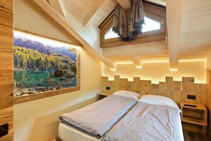 A bedroom with a bed, wooden headboard, bedside tables, a window with a view of mountains and a lake, and a skylight.