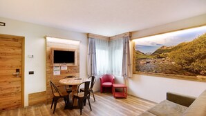 Een kamer met een houten vloer, een aan de muur bevestigde televisie, een eettafel met stoelen, een rode fauteuil, een bank en een groot raam met uitzicht op een landschap.