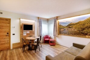 Une pièce avec un plancher en bois, un téléviseur fixé au mur, une table à manger avec des chaises, une chaise rouge, un canapé et une grande fenêtre donnant sur un paysage.