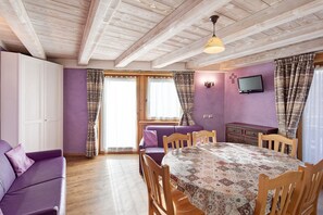 A room with a wooden ceiling, a purple sofa, a dining table with chairs, a television on the wall, and a sliding door with curtains.