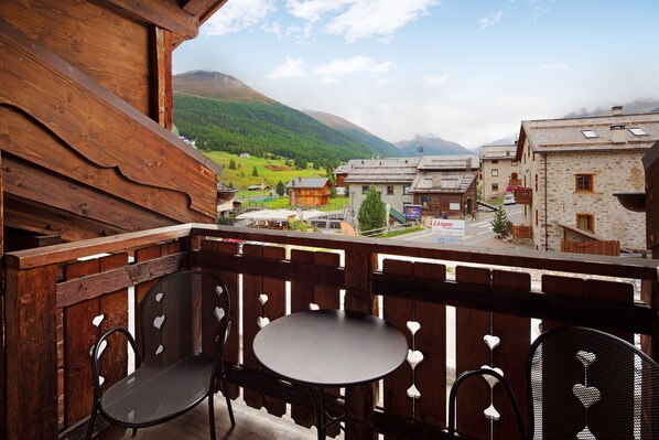 Un balcon en bois, agrémenté de deux chaises et d’une table, d’où l’on peut admirer un village aux bâtiments traditionnels, avec une montagne en toile de fond.