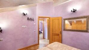 Quarto com porta de madeira, cama com roupa de cama estampada, espelho fixado na parede e corredor que dá para outro cômodo.