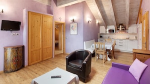 Quarto com teto de madeira, televisão na parede, sofá, poltrona, mesa de jantar com cadeiras e cozinha.