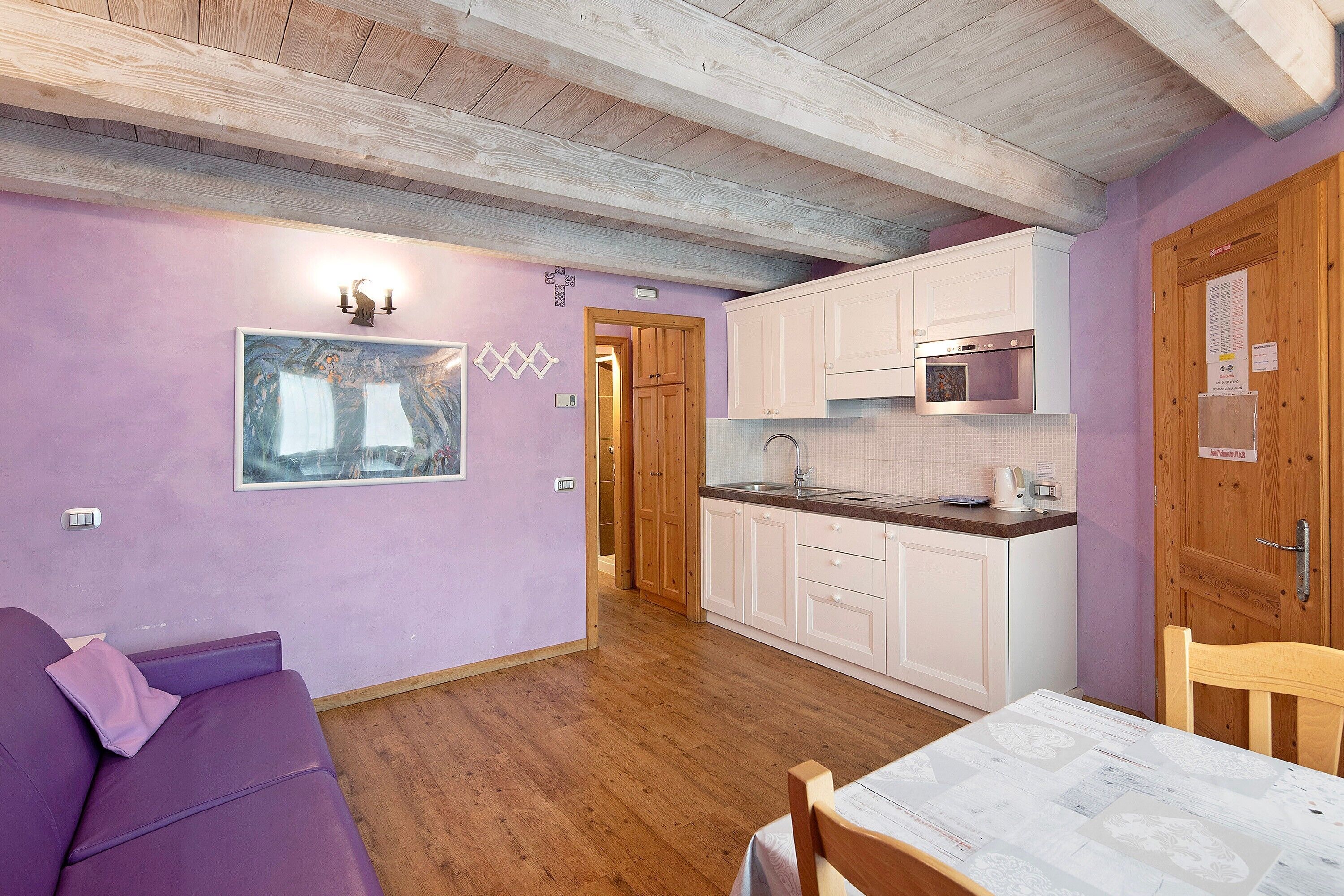 A room with a purple wall, wooden ceiling, a sofa, a table, and a kitchen area with white cabinets and a microwave.