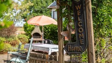 Une clôture en bois ornée d’une pancarte « BIENVENUE CHEZ VOUS », un coin salon extérieur rustique avec une table et des chaises, et un four en briques en arrière-plan.
