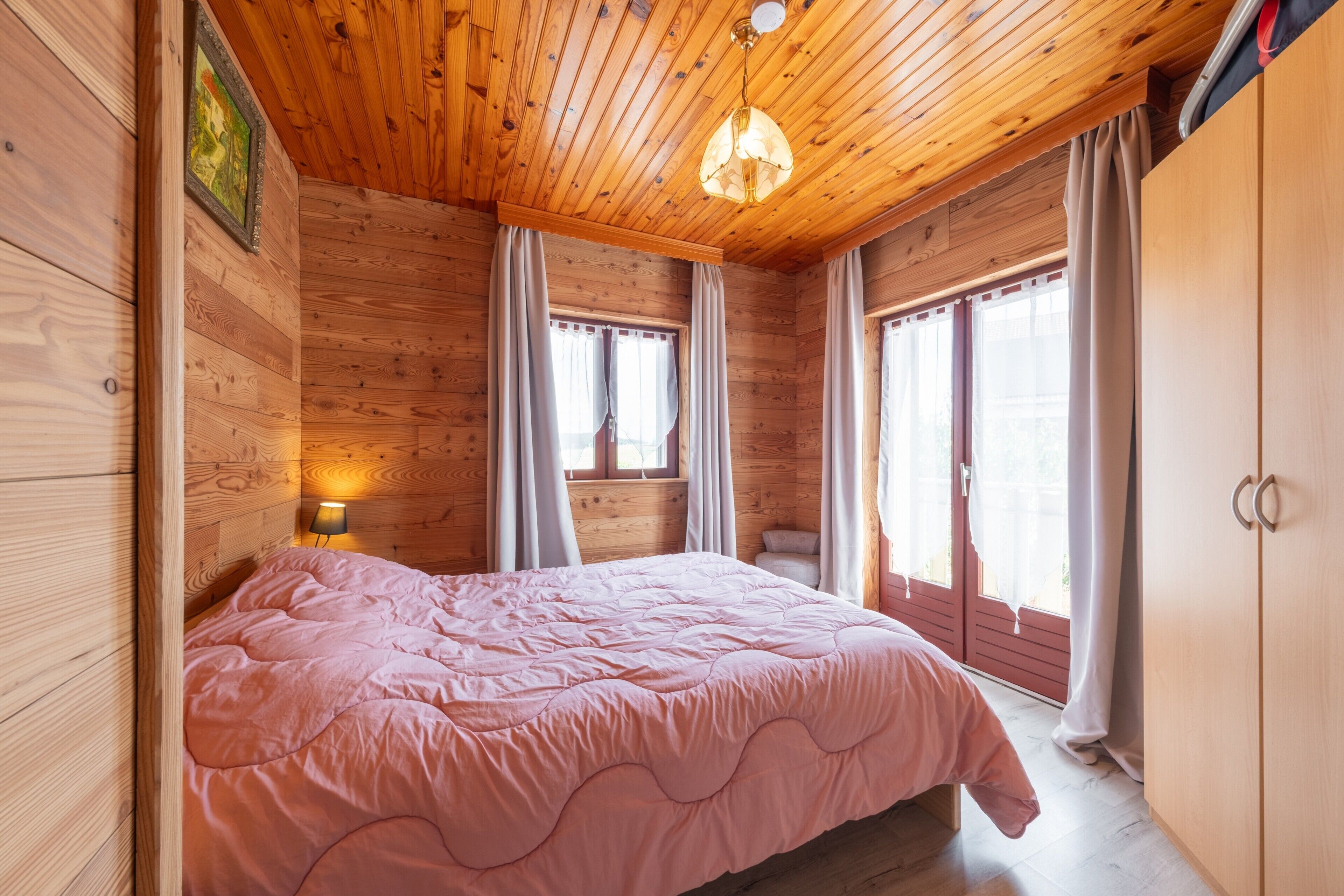 Un dormitorio con techo de madera, una cama con colcha rosa, dos ventanas con cortinas y una mesita de noche con lámpara.