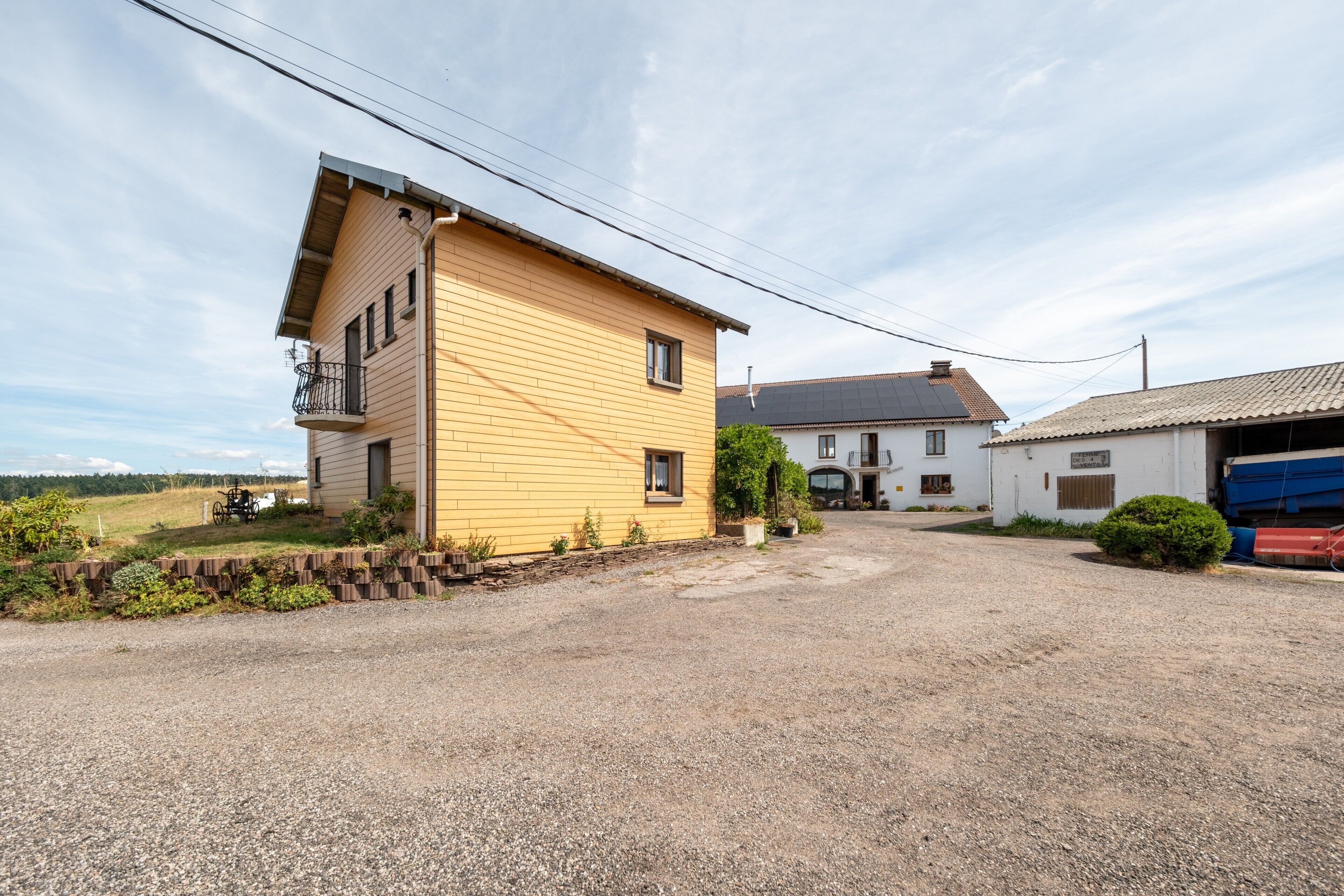 A yellow building with a balcony, a gravel driveway, and a white building with a garage in the background.