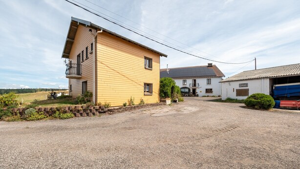 A yellow building with a balcony, a gravel driveway, and a white building with a garage in the background.