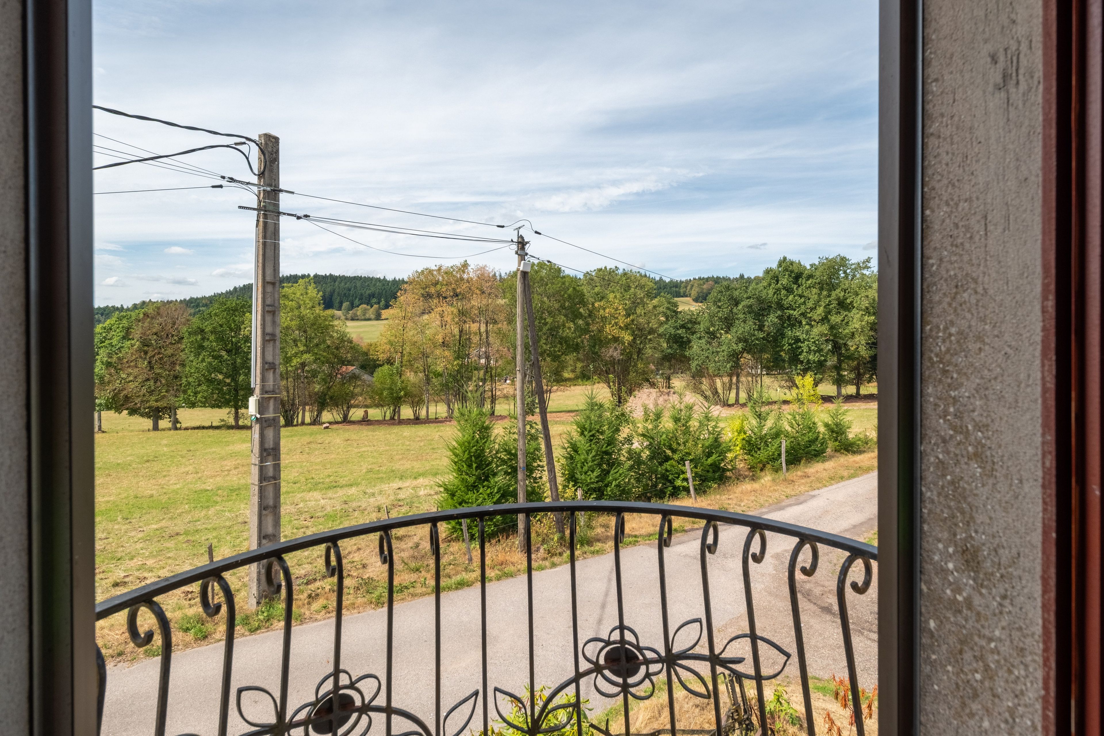A view from a balcony with a metal railing, overlooking a rural landscape with trees and a clear sky.