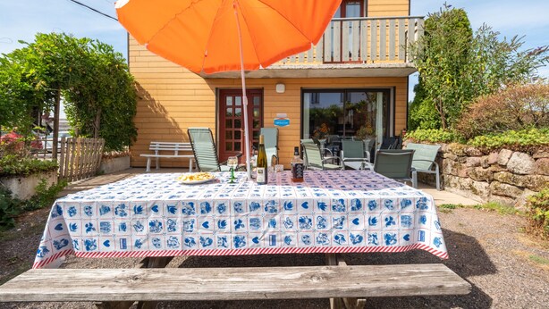 An outdoor dining area with a table set for two, a wooden bench, and a large orange umbrella.