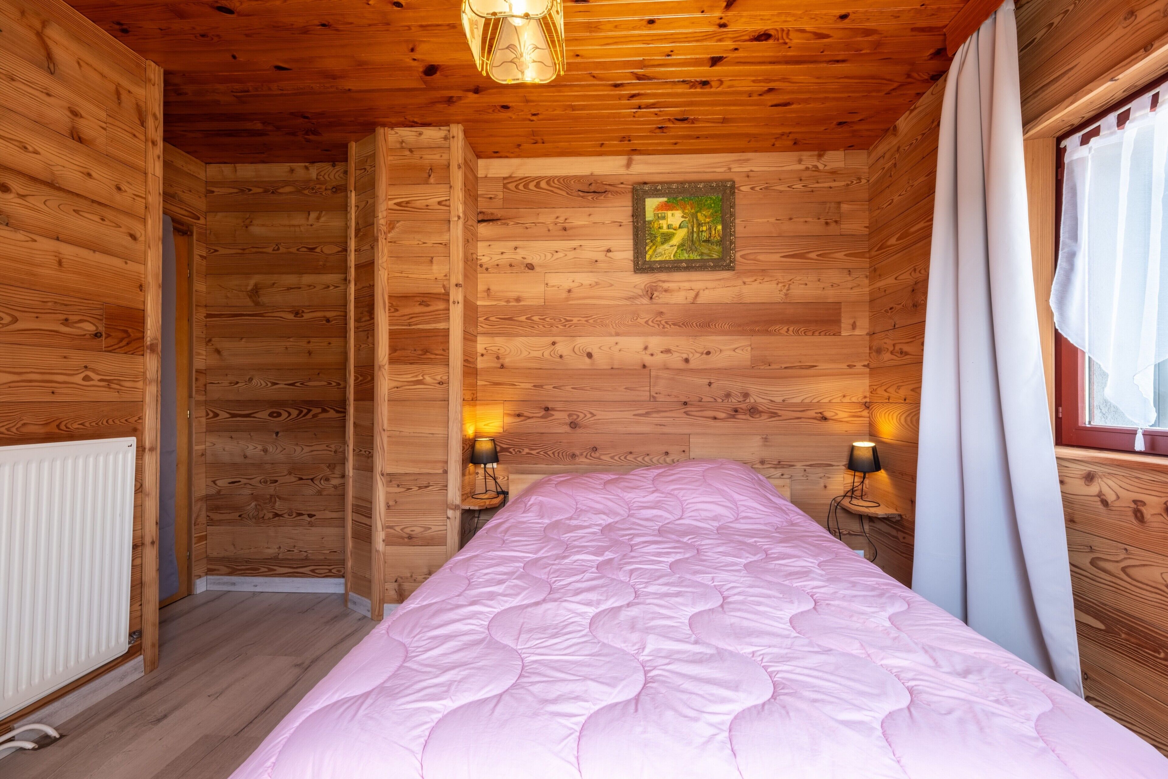 Een slaapkamer in een houten chalet met een bed, twee bedlampjes, een radiator en een raam met gordijnen.