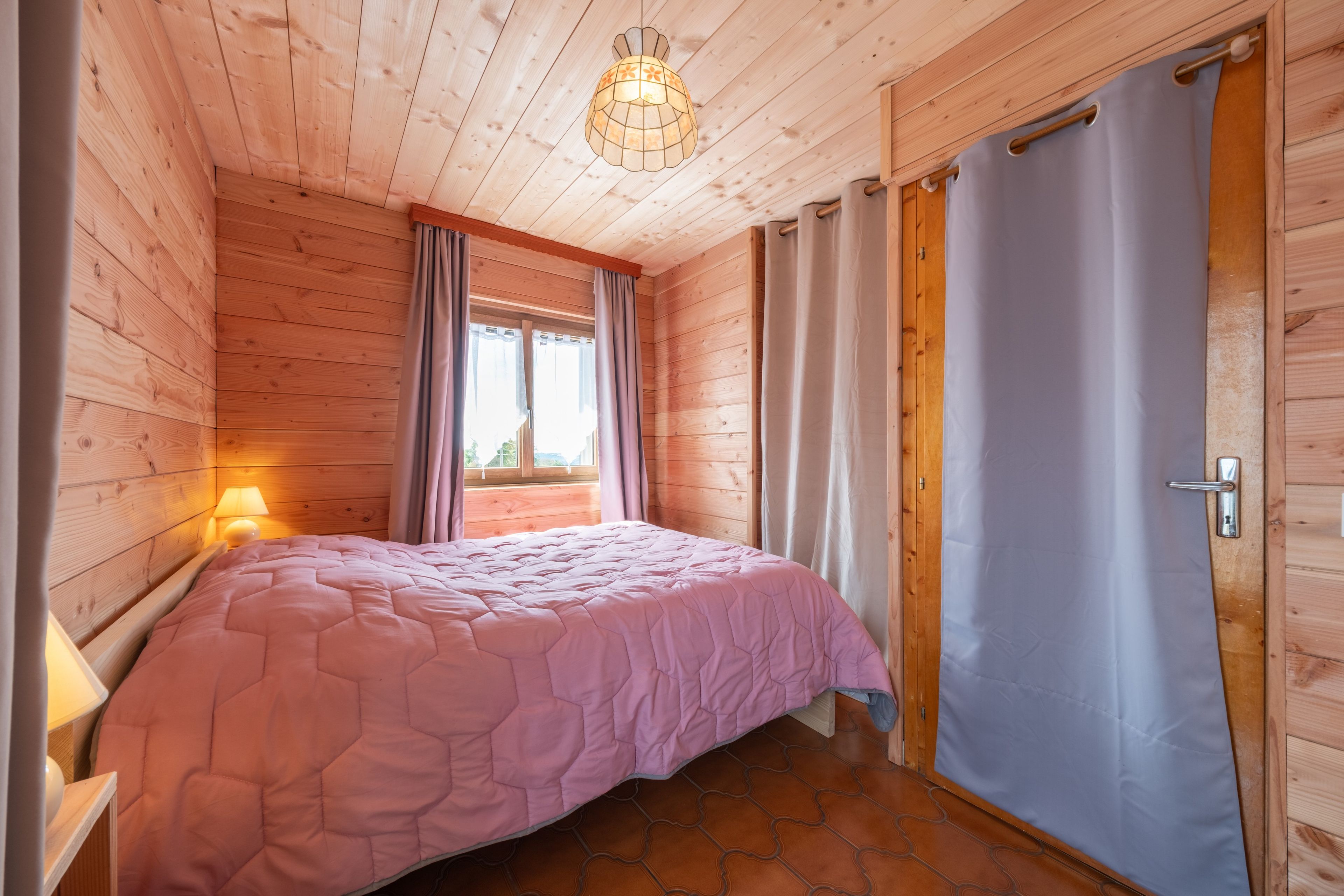 Quarto com paredes de madeira, uma cama com colcha rosa, uma janela com cortinas e uma porta.