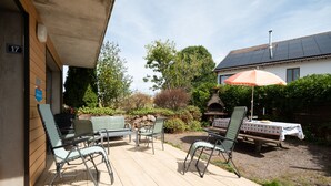 An outdoor patio with seating and a table, surrounded by greenery and a building in the background.