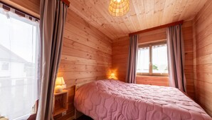 A bedroom with wooden walls, a bed with a pink quilt, a nightstand with a lamp, and a window with curtains.