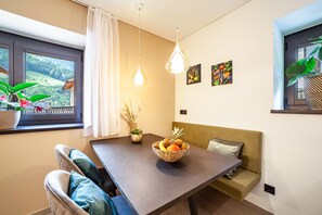 A dining area with a table, chairs, a bowl of fruit, hanging lights, a window with a view, and wall art.
