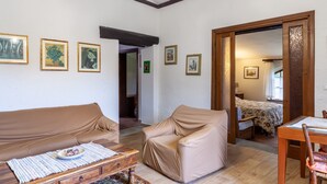 Un salon chaleureux avec un canapé, un fauteuil à oreilles, une table basse en bois et une table à manger avec des chaises. Les murs sont ornés de tableaux encadrés, et une porte donne accès à une autre pièce.