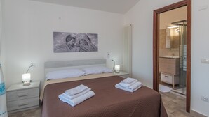 Una camera da letto con un letto, comodini, una lampada da comodino, un'opera d'arte appesa al muro, un bagno con uno specchio e una porta in legno.