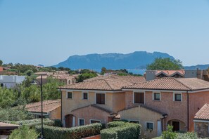 A residential area with houses featuring terracotta roofs and stucco walls, surrounded by greenery and mountains in the background.